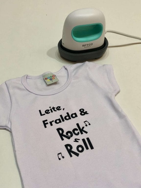 Body de bebê personalizado (roupa infantil) branco, personalizado com a frase na cor preta "Leite, Fralda & Rock n Roll". Em cima da roupa uma mini prensa térmica branca e azul. 